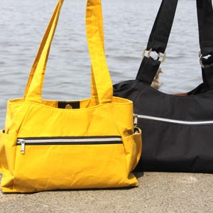 Könnte beinhalten: Zwei Tragetaschen, eine gelbe und eine schwarze, stehen nebeneinander. Die gelbe Tasche hat einen schwarz-weißen Streifen und eine Reißverschlusstasche. Die schwarze Tasche hat braune Lederakzente. Beide haben Schultergurte.