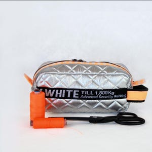 Può includere: Pochette cosmetica argentata trapuntata con dettagli arancioni e una fascia nera con la scritta "WHITE TILL 1,800Kg Advanced Security Webbing". Due rocchetti di filo arancione e forbici nere sono accanto alla borsa.