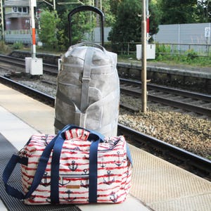 May include: Two travel bags on a train platform. A large, light gray rolling suitcase stands upright. In front, a smaller duffel bag features a white background with red stripes and anchor designs, and has blue straps.