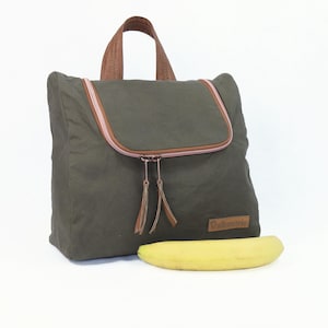 Könnte beinhalten: Olivgrüner Rucksack mit braunem Ledergriff und -besatz. Der Rucksack hat einen rosa Reißverschluss und ein kleines braunes Lederetikett. Eine gelbe Banane liegt vor dem Rucksack.