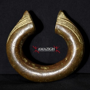 May include: A bronze-colored, open-ended cuff bracelet. The bracelet has a rounded, thick band with a textured design on the open ends. The ends have a lighter, cream-colored pattern. The word "AMAZIGH" is visible in red.