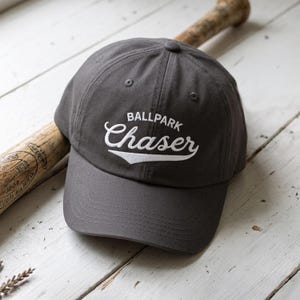 May include: A charcoal gray baseball cap featuring the embroidered white text BALLPARK Chaser with a swoosh underline. The cap has a structured crown, a curved brim, and a button on top, positioned next to a wooden baseball bat.