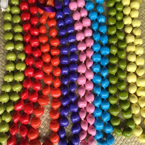 May include: A close-up of eight colorful leis made of seed pods. The leis are arranged in a row, with the colors ranging from green to yellow to orange to red to purple to pink to blue to turquoise.
