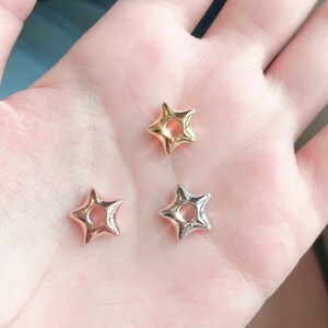 Puede incluir: Tres colgantes en forma de estrella en tonos dorados, oro rosa y plateado. Cada colgante tiene un centro hueco y una superficie lisa y pulida. Los colgantes son pequeños y delicados, probablemente para la fabricación de joyas o manualidades.