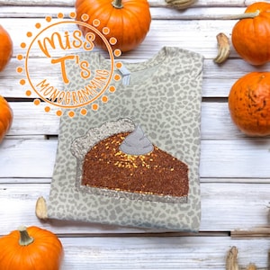 May include: A light brown t-shirt with a leopard print design. The shirt features a sparkly orange and brown pumpkin pie slice with a white whipped cream topping. The text "Miss T's Monogramming" is in a circle on the top left corner of the shirt.