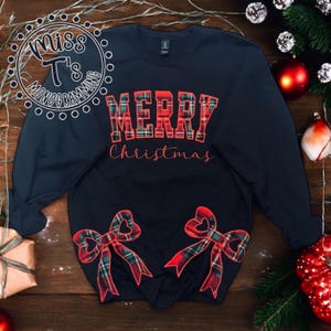 May include: A black long-sleeved sweatshirt with the text "Merry Christmas" in red and white plaid. The sweatshirt has two red and green plaid bows on the bottom.
