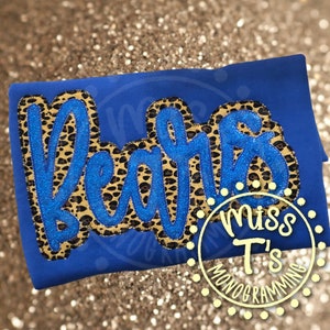 May include: A royal blue t-shirt with the word "Bears" in a blue glitter script, outlined with a leopard print pattern. The shirt also features a circular logo with the text "Miss T's Monogramming".