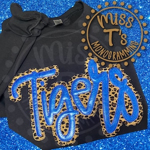May include: Black t-shirt with a blue and brown "Tigers" design. The design has a leopard print pattern. The shirt is on a blue sparkly background. The text "Miss T's Monogramming" is in a circle in the top right corner.