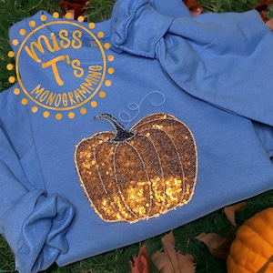 Puede incluir: Una sudadera azul con un diseño de calabaza de lentejuelas doradas. La sudadera tiene el texto "Miss T's Monogramming" en un círculo en la esquina superior izquierda.