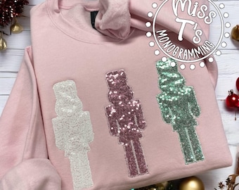 May include: Pink sweatshirt with three sequined nutcracker designs. The nutcrackers are white, pink, and green.