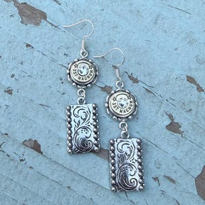 May include: A pair of silver-toned drop earrings featuring a circular 357 Magnum cartridge casing top with a central clear crystal, connected to a rectangular pendant with an embossed floral scroll pattern and a beaded border edge.