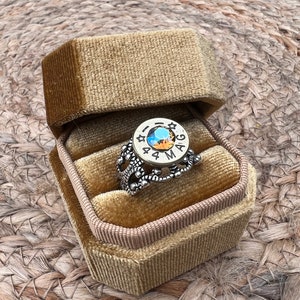 May include: A silver-toned ring with a central, multi-colored gemstone is displayed inside a gold velvet ring box. The ring features intricate filigree detailing and text around the gemstone. The ring box is open, revealing the ring against a textured background.