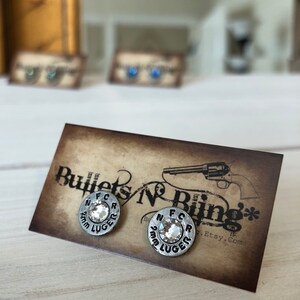 May include: Silver-toned stud earrings with a clear, faceted center stone. The earrings are circular with the text "N F C R 9mm LUGER" around the edge. The earrings are displayed on a brown card with the text "Bullets N Bling".