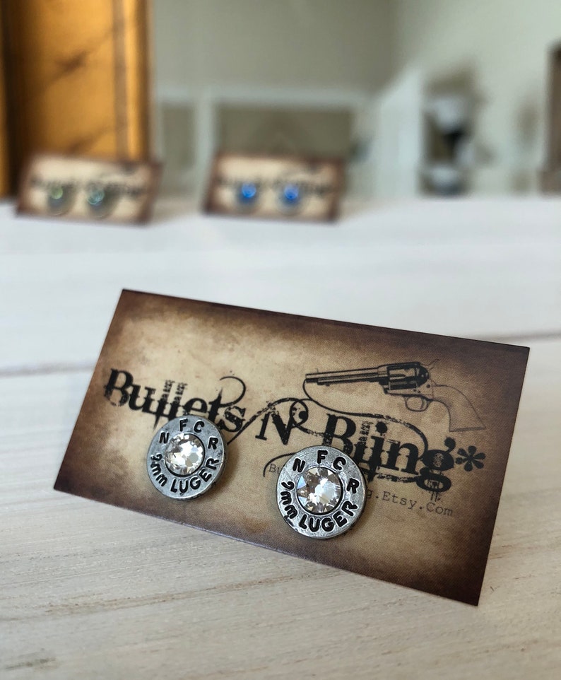 May include: Silver-toned stud earrings with a clear, faceted center stone. The earrings are circular with the text "N F C R 9mm LUGER" around the edge. The earrings are displayed on a brown card with the text "Bullets N Bling".