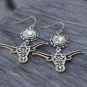K&ouml;nnte beinhalten: Ein Paar goldfarbene Ohrringe mit einem Longhorn-Design. Jeder Ohrring hat eine silberfarbene Oberseite mit einem Kristall und dem Text "R-P LUGER". Die Ohrringe h&auml;ngen an goldfarbenen Haken.