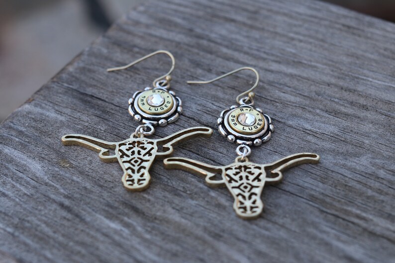 K&ouml;nnte beinhalten: Ein Paar goldfarbene Ohrringe mit einem Longhorn-Design. Jeder Ohrring hat eine silberfarbene Oberseite mit einem Kristall und dem Text "R-P LUGER". Die Ohrringe h&auml;ngen an goldfarbenen Haken.