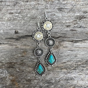 May include: Silver-toned dangle earrings with a Western theme. Each earring features a bullet casing with the text "SPEER 9mm LUGER", decorative floral accents, and a teardrop-shaped turquoise stone. The earrings hang from silver hooks.
