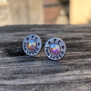 May include: A pair of silver-toned stud earrings. Each earring is a small, circular design with the text "N F C R 9mm LUGER" stamped around the edge. The center of each earring features a shimmering, iridescent crystal. The earrings are displayed on a weathered wooden surface.