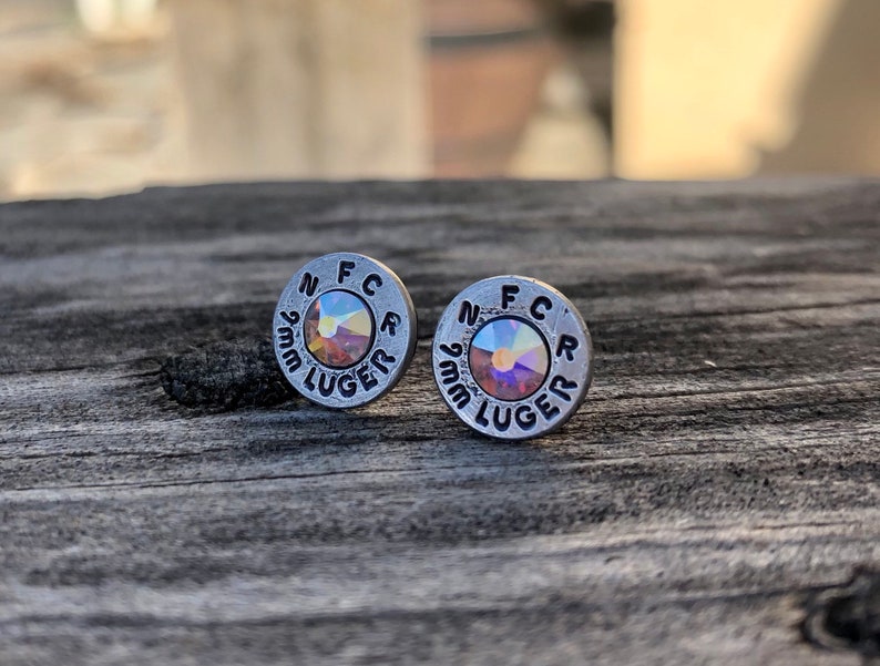 May include: A pair of silver-toned stud earrings. Each earring is a small, circular design with the text "N F C R 9mm LUGER" stamped around the edge. The center of each earring features a shimmering, iridescent crystal. The earrings are displayed on a weathered wooden surface.