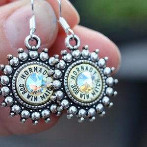 May include: A pair of silver-toned earrings with a unique design. Each earring features a central iridescent gem surrounded by a sunburst pattern of small silver beads. The center has the text "HORNADY 308 WIN MATCH".