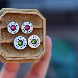 May include: Four stud earrings in a velvet jewelry box. Two pairs of silver-toned earrings feature a red or green gemstone center. The earrings are made from bullet casings with the markings "CC/R 38 SPL" and "FC .45 AUTO".