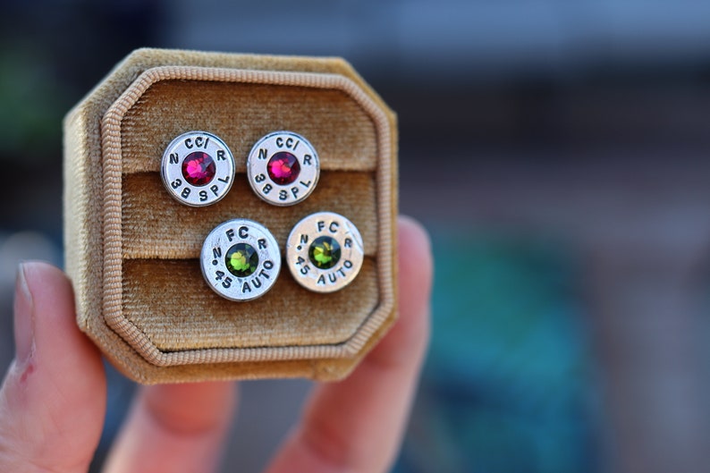 May include: Four stud earrings in a velvet jewelry box. Two pairs of silver-toned earrings feature a red or green gemstone center. The earrings are made from bullet casings with the markings "CC/R 38 SPL" and "FC .45 AUTO".