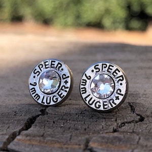 May include: A pair of silver-toned stud earrings. Each earring is circular with the text "SPEER 9mm LUGER +p" around a central clear crystal. The earrings are set against a blurred wooden background.