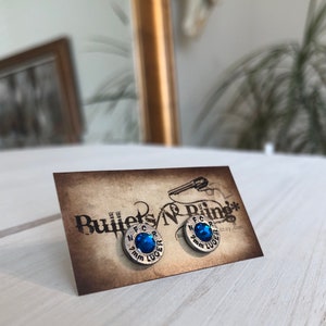 May include: Silver-toned stud earrings with a blue gemstone center, set on a brown card. The earrings are shaped like bullet casings, with the text "9mm LUGER" and "N.F.C.R." visible. The card has the text "Bullets N' Bling".