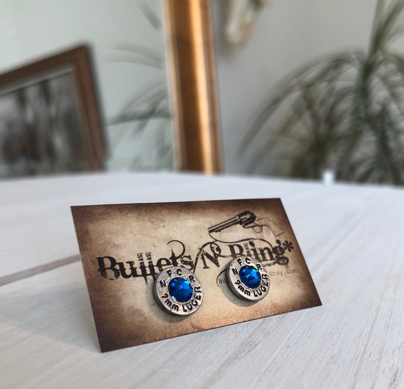 May include: Silver-toned stud earrings with a blue gemstone center, set on a brown card. The earrings are shaped like bullet casings, with the text "9mm LUGER" and "N.F.C.R." visible. The card has the text "Bullets N' Bling".