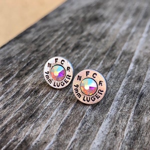 May include: A pair of silver-toned stud earrings. Each earring is circular with the text "FCR" and "9mm LUGER" engraved around the edge. The center of each earring features a shimmering, iridescent crystal.