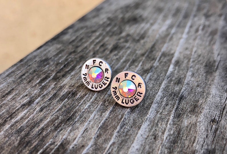 May include: A pair of silver-toned stud earrings. Each earring is circular with the text "FCR" and "9mm LUGER" engraved around the edge. The center of each earring features a shimmering, iridescent crystal.