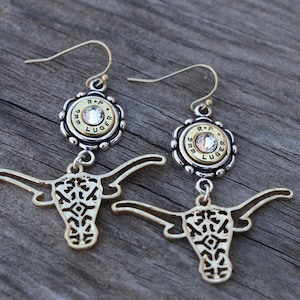 K&ouml;nnte beinhalten: Ein Paar goldfarbene Ohrringe mit Longhorn-Design. Jeder Ohrring hat eine silberfarbene Fassung mit einem klaren Edelstein und einer Patronenh&uuml;lse. Die Ohrringe h&auml;ngen an einem silberfarbenen Haken.