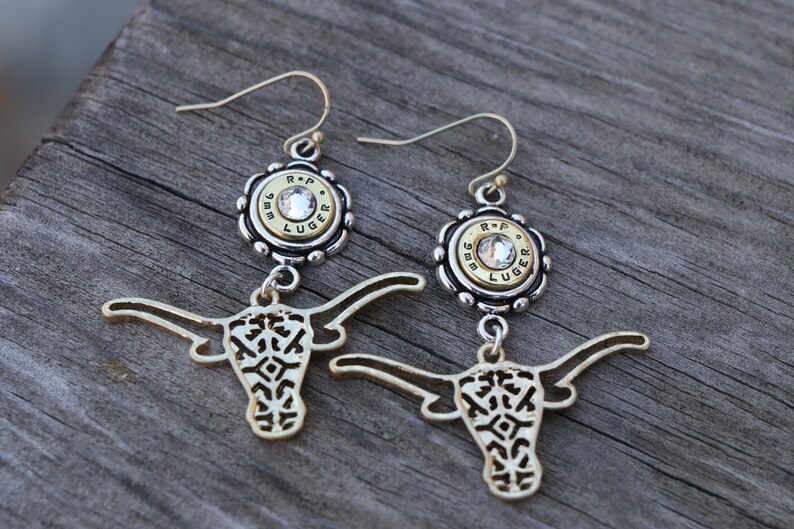 K&ouml;nnte beinhalten: Ein Paar goldfarbene Ohrringe mit Longhorn-Design. Jeder Ohrring hat eine silberfarbene Fassung mit einem klaren Edelstein und einer Patronenh&uuml;lse. Die Ohrringe h&auml;ngen an einem silberfarbenen Haken.