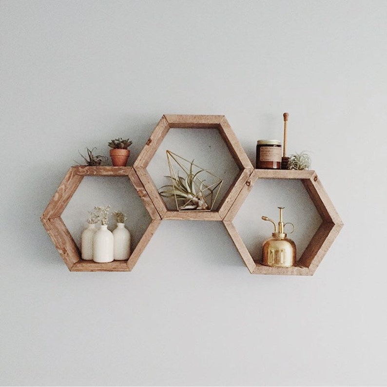Puede incluir: Tres estantes hexagonales de madera montados en una pared. Los estantes est&aacute;n dispuestos en forma de panal. El estante superior tiene un peque&ntilde;o frasco de vidrio y un palo de madera. El estante del medio tiene una peque&ntilde;a planta en maceta y una escultura geom&eacute;trica dorada. El estante inferior tiene tres peque&ntilde;as botellas blancas y una regadera dorada.