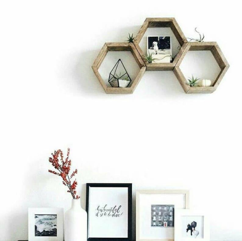 Puede incluir: Tres estantes hexagonales de madera montados en una pared blanca. Los estantes est&aacute;n dispuestos en forma escalonada y presentan peque&ntilde;as plantas y una foto enmarcada. Los estantes son de color marr&oacute;n claro y tienen un acabado r&uacute;stico.