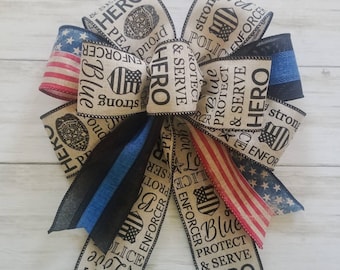 May include: A decorative bow with patriotic themes. The bow features ribbons with the American flag, blue stripes, and text that reads "Blue", "Hero", "Protect & Serve", and "Police".
