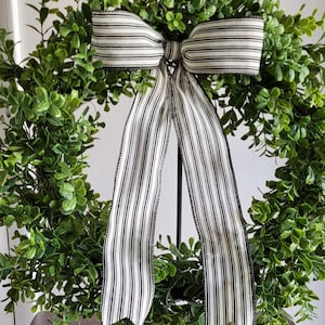 May include: A round, green wreath with lush, leafy foliage. A large bow made of black and white striped fabric is centered at the top, with long, flowing tails. The wreath is a decorative item, suitable for seasonal or year-round display.