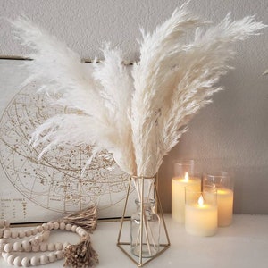 May include: A decorative arrangement featuring fluffy, cream-colored pampas grass in a geometric gold vase. The display includes a string of wooden beads, a framed map, and three glowing pillar candles, creating a warm, inviting aesthetic.