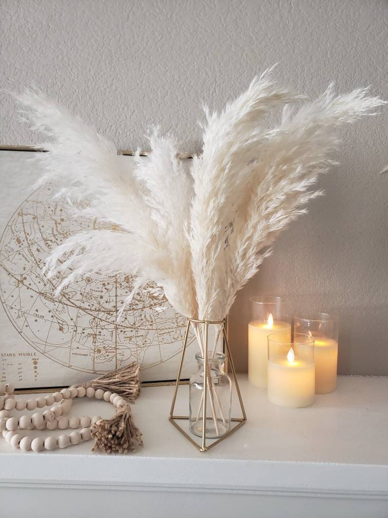 May include: A decorative arrangement featuring fluffy, cream-colored pampas grass in a geometric gold vase. The display includes a string of wooden beads, a framed map, and three glowing pillar candles, creating a warm, inviting aesthetic.