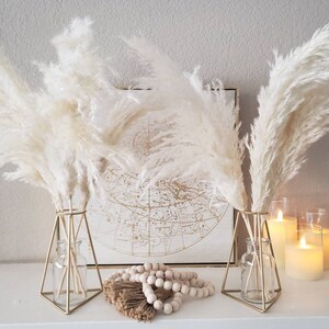 May include: Two gold geometric vases with fluffy, cream-colored pampas grass. A framed map and candles are in the background. Decorative wooden beads and a bundle of dried stems are in front of the vases.