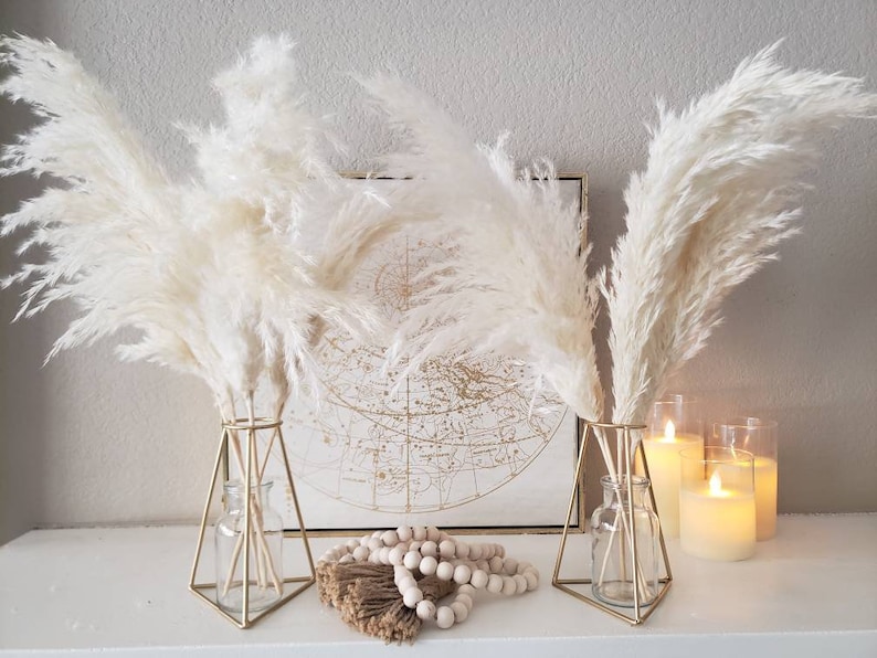 May include: Two gold geometric vases with fluffy, cream-colored pampas grass. A framed map and candles are in the background. Decorative wooden beads and a bundle of dried stems are in front of the vases.