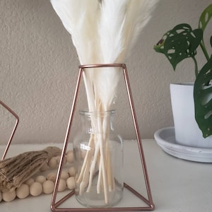May include: White pampas grass in a clear glass vase, sitting on a rose gold triangular stand.