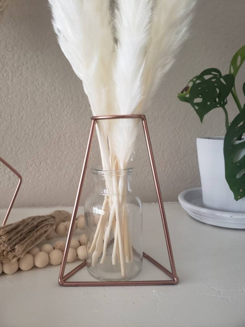 May include: White pampas grass in a clear glass vase, sitting on a rose gold triangular stand.