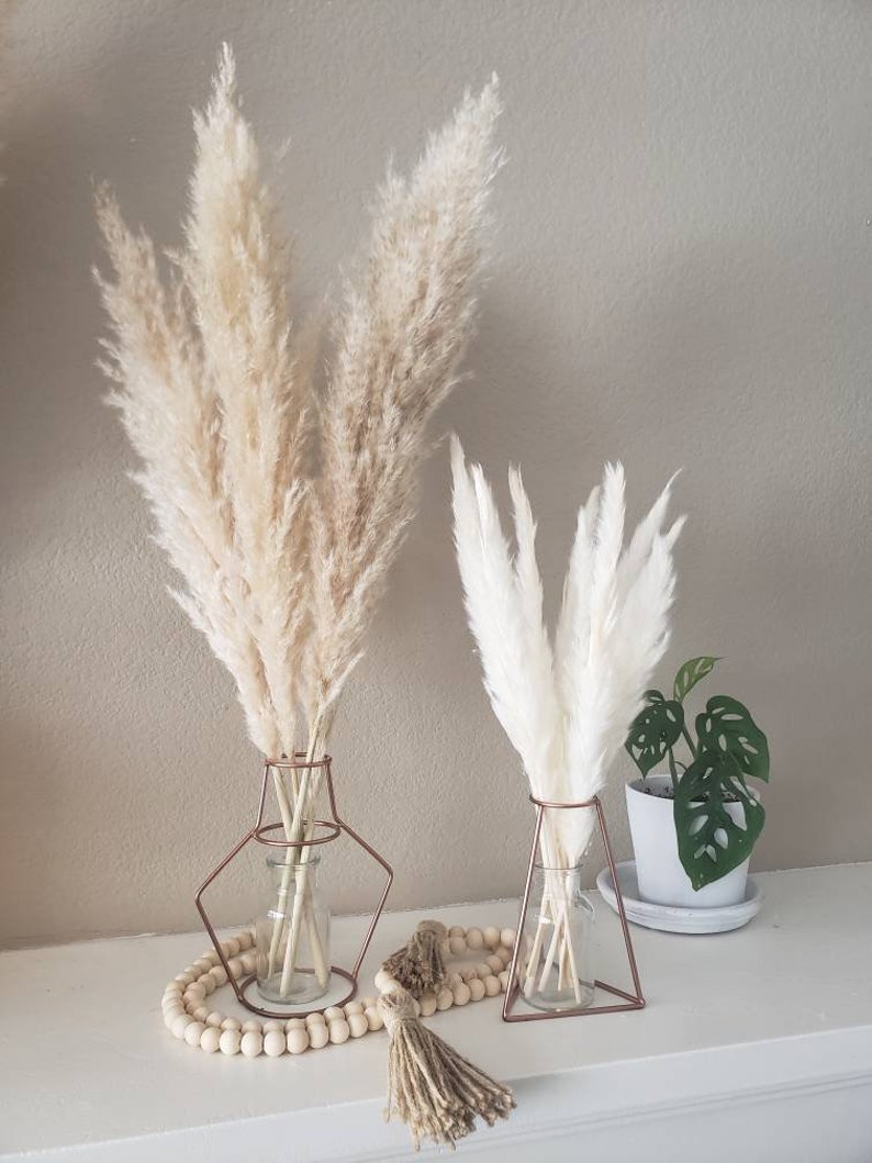 May include: Two pampas grass arrangements in clear glass vases with copper wire frames. The vases are on a white surface with a wooden bead garland and a brown tassel.