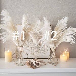 May include: A decorative display featuring two bouquets of fluffy, cream-colored pampas grass in gold geometric vases. The display includes lit candles in glass holders, a string of wooden beads, and a framed map.