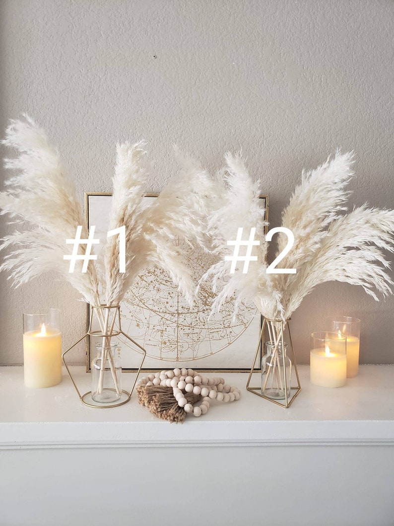 May include: A decorative display featuring two bouquets of fluffy, cream-colored pampas grass in gold geometric vases. The display includes lit candles in glass holders, a string of wooden beads, and a framed map.
