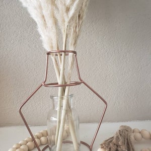 May include: A clear glass vase with a copper wire frame sits on a table with a strand of wooden beads. The vase is filled with dried pampas grass.