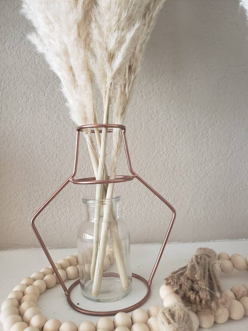 May include: A clear glass vase with a copper wire frame sits on a table with a strand of wooden beads. The vase is filled with dried pampas grass.