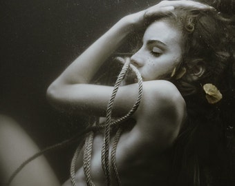 May include: A woman with long dark hair is submerged in water. She is bound with a thick rope and has a leaf near her ear.