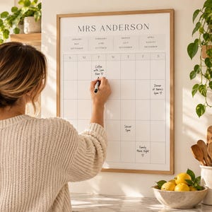 May include: A wall-mounted dry-erase calendar with a light wood frame featuring the text MRS ANDERSON at the top. The white board has a grid layout for days of the week and months of the year, with handwritten notes including Coffee with Sam, Dinner at Nana's, Soccer, and Family Movie Night.